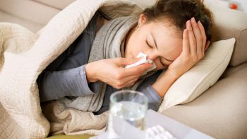 Dame ligt op bed met zakdoek en snuit met haar ene hand haar neus en heeft andere hand op haar voorhoofd